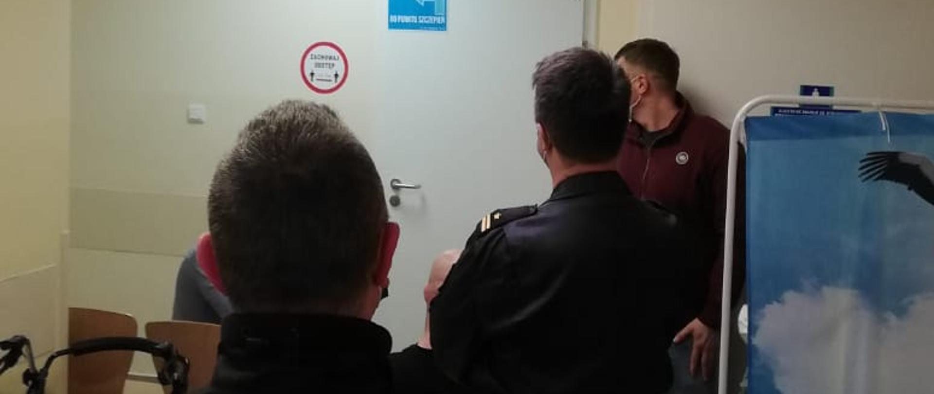 Zdjęcie przedstawia funkcjonariuszy z Komendy Powiatowej PSP w Tucholi (w umundurowaniu koszarowym oraz w ubraniu cywilnym), czekających w kolejce do punktu szczepień przeciwko COVID-19.