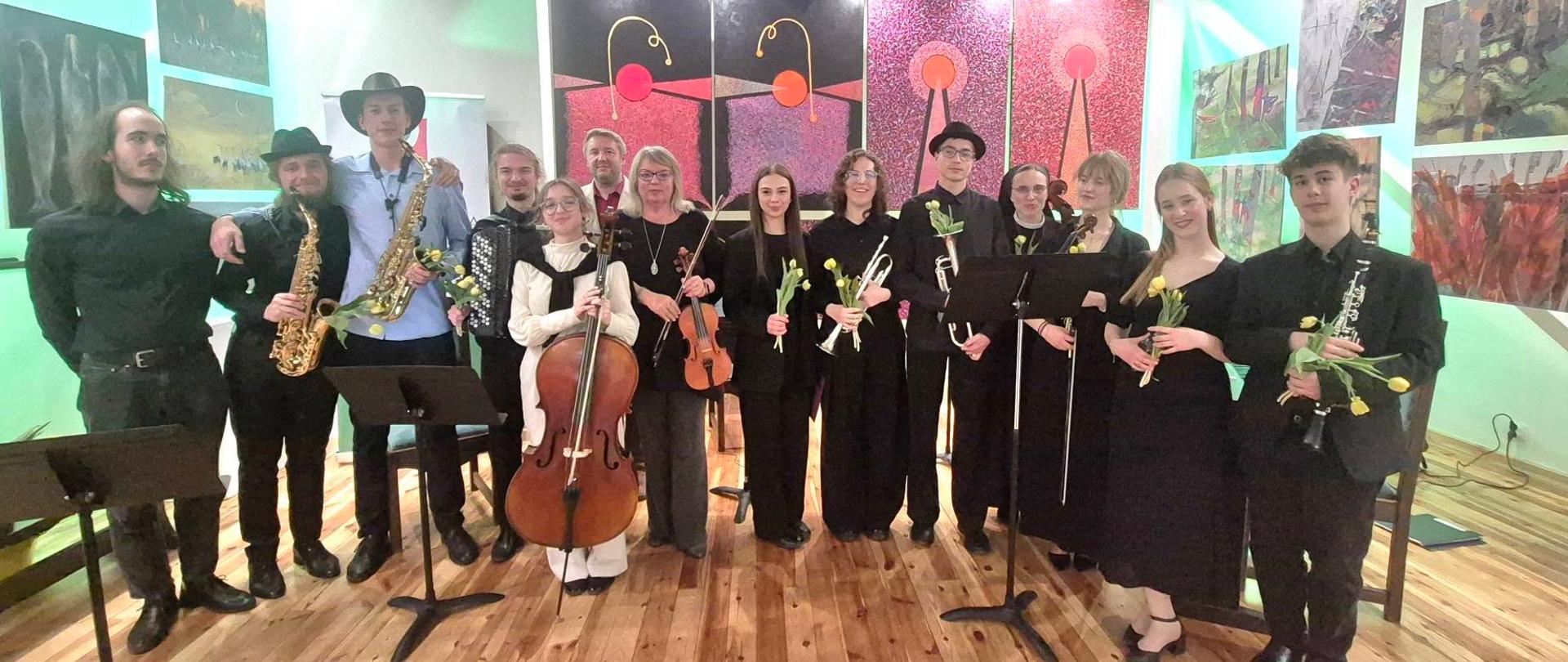 Młodzież i nauczyciele w uroczystych strojach z instrumentami. Na ścianach galeria z obrazami