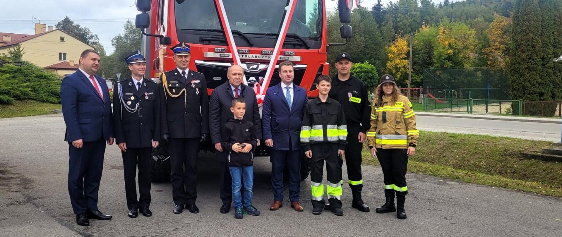 Zdjęcie zrobione na zewnątrz podczas uroczystości. Szeregu stoją uczestnicy uroczystości, wśród nich oficer Państwowej Straży Pożarnej w stopnia starszego brygadiera. Za nimi, przepasany biało-czerwoną szarfą, samochód ratowniczo-gaśniczy.