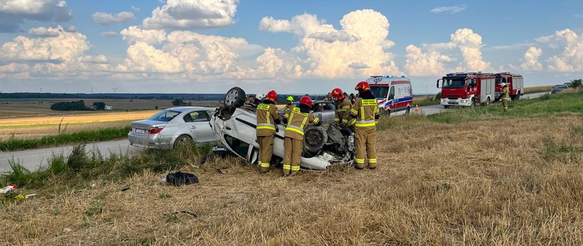 Zdjęcie przedstawia rozbity samochód osobowy znajdujący się na dachu poza jezdnią. Przy uszkodzonym pojeździe widać strażaków w umundurowaniu specjalnym, którzy odłączają zasilanie elektryczne w pojeździe. W tle zaparkowane na drodze pojazdy karetki pogotowia oraz samochody ratowniczo-gaśnicze. 