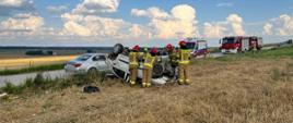 Zdjęcie przedstawia rozbity samochód osobowy znajdujący się na dachu poza jezdnią. Przy uszkodzonym pojeździe widać strażaków w umundurowaniu specjalnym, którzy odłączają zasilanie elektryczne w pojeździe. W tle zaparkowane na drodze pojazdy karetki pogotowia oraz samochody ratowniczo-gaśnicze. 