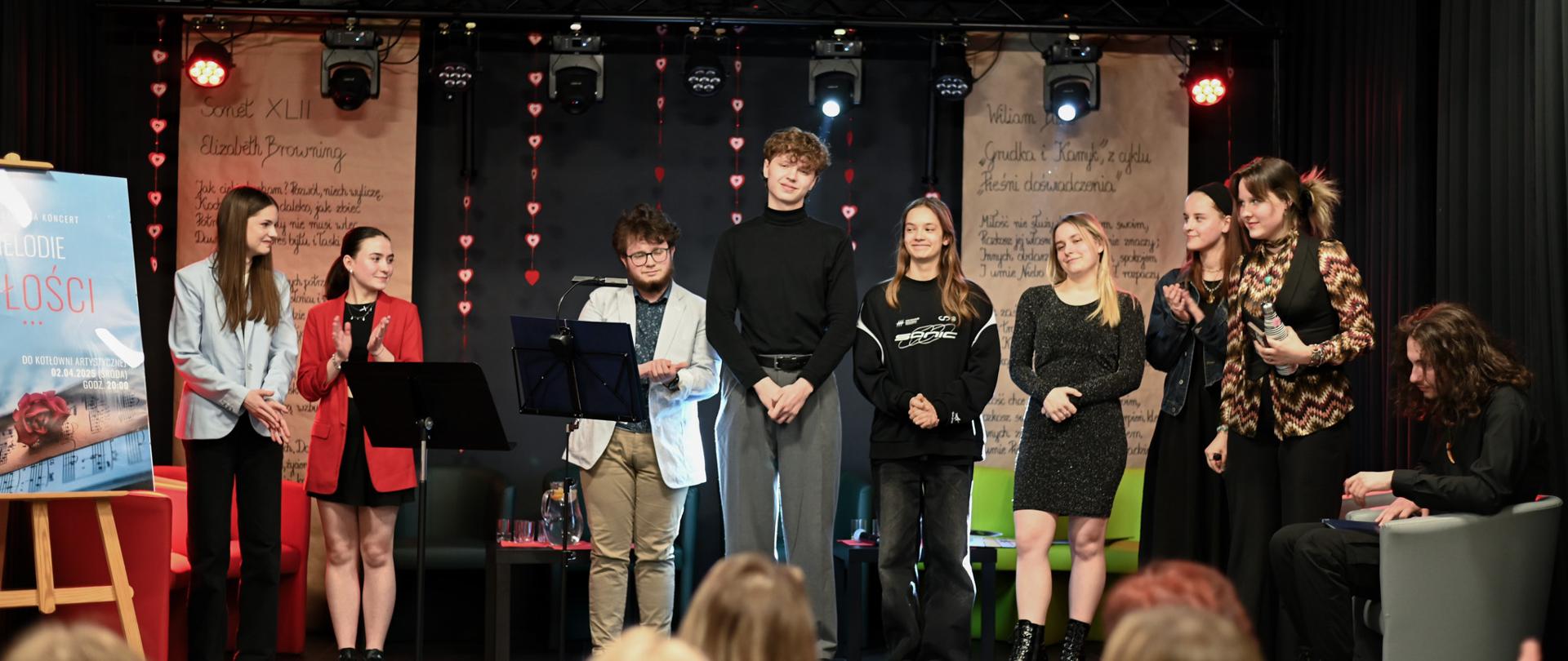 Na scenie stoi kilkoro młodych ludzi, za nimi są stoliki z napojami w dzbankach i fotele. W tle widać dwa duże zwisające z sufitu arkusze szarego papieru z tekstami wierszy Wiliama Blake'a i Elizabeth Browning. Pomiędzy nimi zwisają czerwono-białe serduszka na paskach. Na górze listwa z reflektorami. w pierwszym planie widać tył głów publiczności. Po lewej stronie na sztaludze stoi plakat z napisem MELODIE MIŁOŚCI. W pierwszym planie widać tyły głów publiczności.