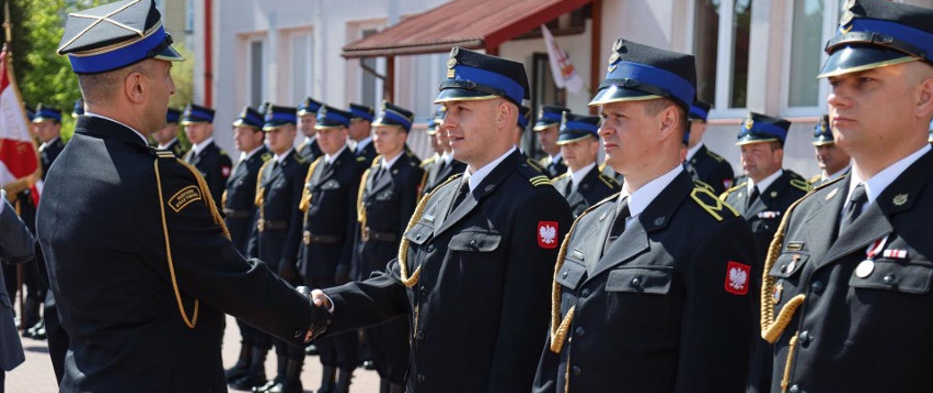 Zdjęcie zrobione na zewnątrz podczas uroczystości. Oficer Państwowej Straży Pożarnej w stopniu starszego brygadiera gratuluje wyróżnienia strażakowi, który stoi w szeregu z innymi strażakami. Strażacy podają sobie dłonie. Z tyłu stoi kompania honorowa złożona z funkcjonariuszy Państwowej Straży Pożarnej. 