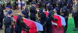 Strażacy z kompani reprezentacyjnej stoją obok trumien i przygotowują flagę Polski do zwinięcia obok stoją uczestnicy uroczystości pogrzebowej. 