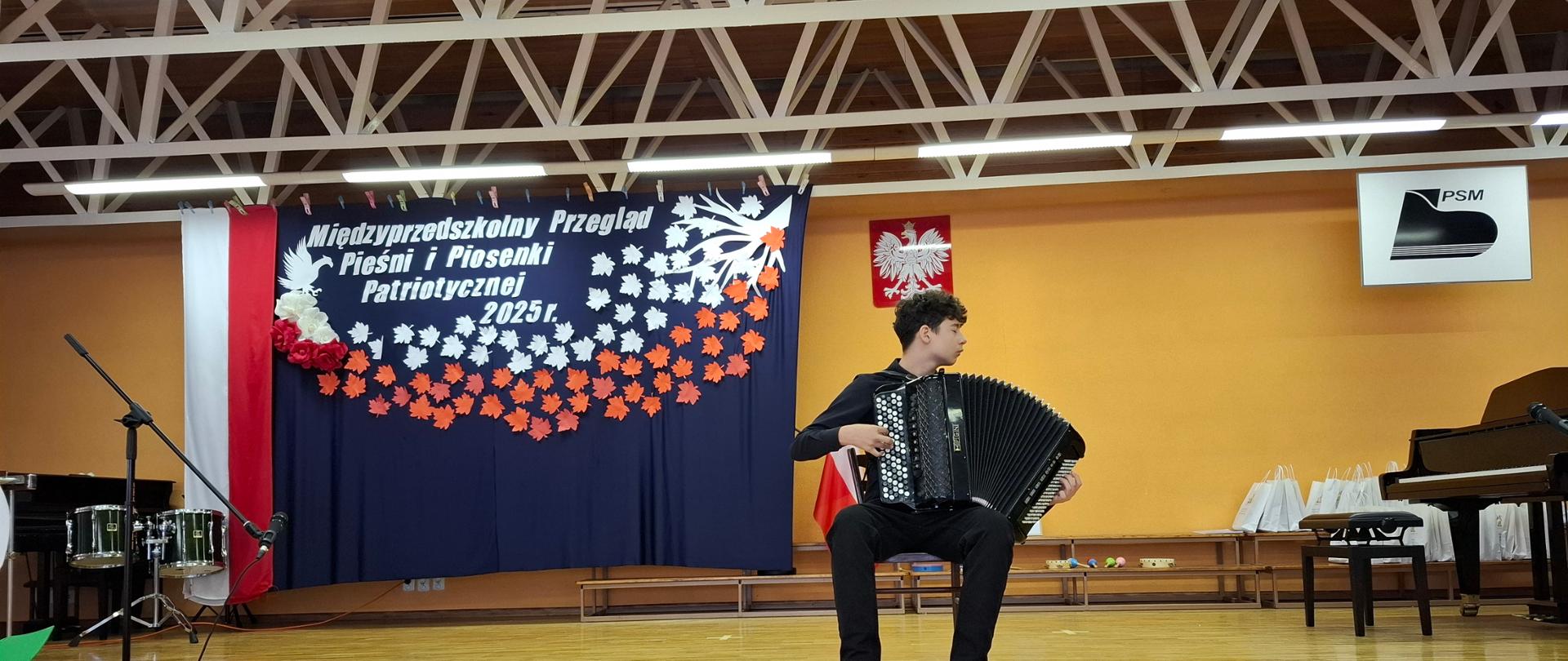 Uczeń gra na instrumencie muzycznym. W tle instrumenty muzyczne, dekoracja biało-czerwona.