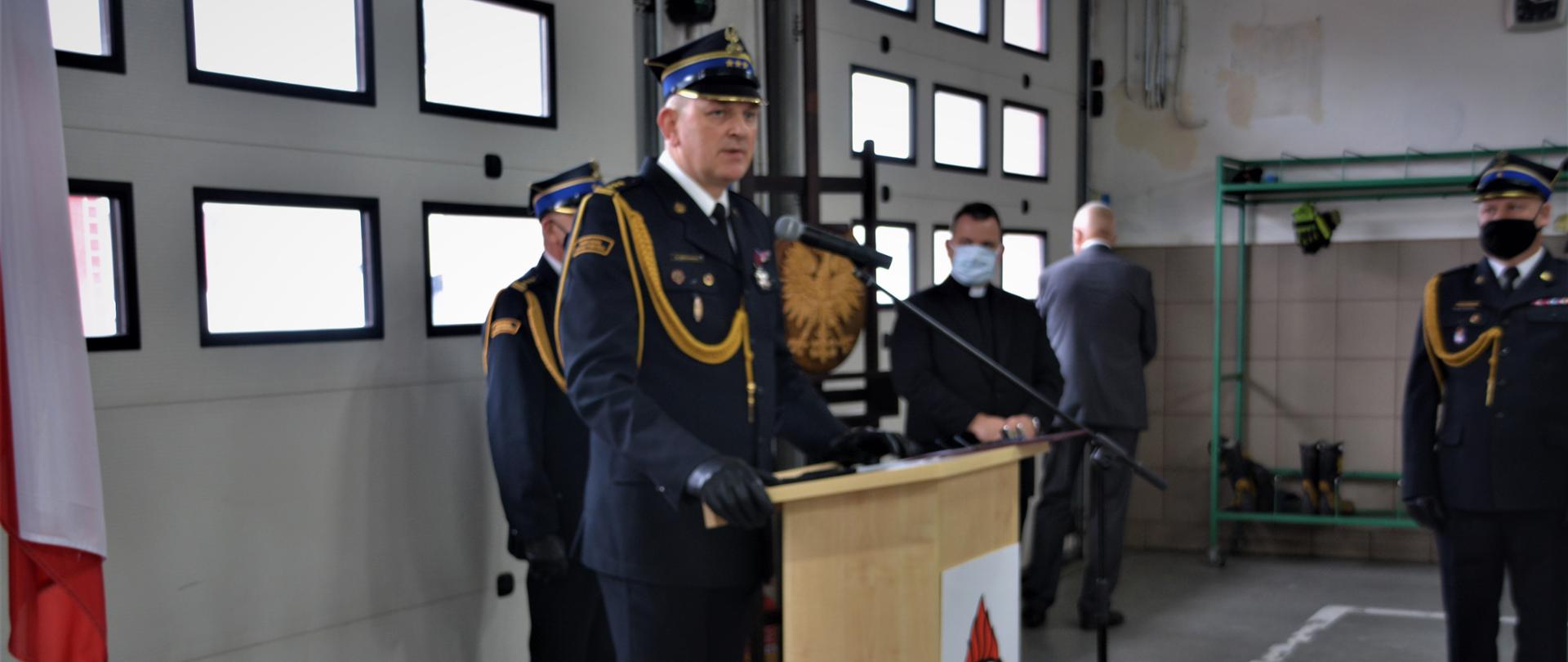 1. Na pierwszym planie w mundurze wyjściowym Zastępca Kujawsko-Pomorskiego Komendanta Wojewódzkiego Państwowej Straży Pożarnej przemawiający przy mównicy. Na zdjęciu widać również w mundurach wyjściowych Komendanta Powiatowego PSP w Tucholi i Dowódcę uroczystości oraz księdza kapelana wojewódzkiego strażaków. Wszyscy stoją w garażu JRG w Tucholi na tle bram garażowych. Na zdjęciu widać flagi państwowe i godło.