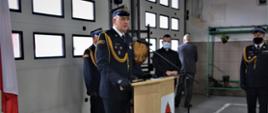 1. Na pierwszym planie w mundurze wyjściowym Zastępca Kujawsko-Pomorskiego Komendanta Wojewódzkiego Państwowej Straży Pożarnej przemawiający przy mównicy. Na zdjęciu widać również w mundurach wyjściowych Komendanta Powiatowego PSP w Tucholi i Dowódcę uroczystości oraz księdza kapelana wojewódzkiego strażaków. Wszyscy stoją w garażu JRG w Tucholi na tle bram garażowych. Na zdjęciu widać flagi państwowe i godło.