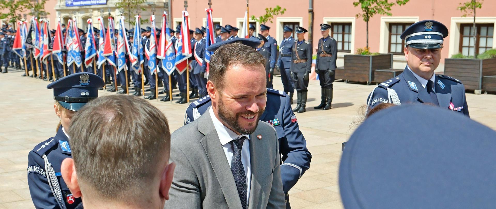 Wojewoda ściska rękę osobie. Za nim kobieta i dwóch mężczyzn w mundurach policji. W tle ustawieni w szeregu policjanci oraz poczty sztandarowe.