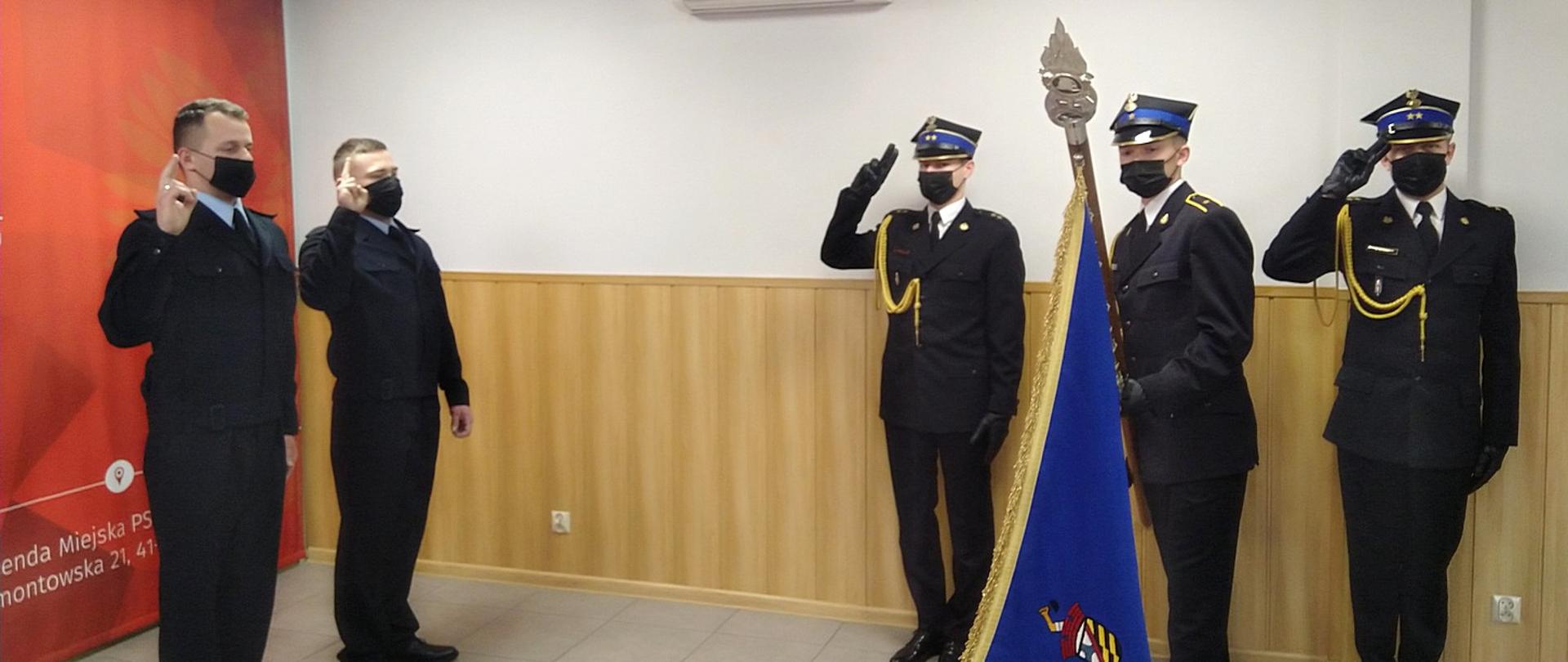 Po lewej stronie zdjęcia ślubujący strażacy stojący naprzeciwko sztandaru. Po prawej stronie zdjęcia widoczny poczet sztandarowy Komendy Miejskiej PSP w Sosnowcu.