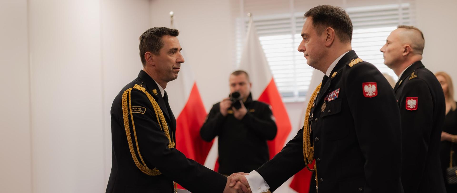 Zdjęcie przedstawia dwóch wysokich rangą oficerów Państwowej Straży Pożarnej uściskujących sobie dłonie podczas oficjalnej uroczystości. Obaj są ubrani w galowe mundury z odznaczeniami, złotymi sznurami naramiennymi i naszywkami z godłem Polski na ramionach. W tle widać dwie polskie flagi, fotografa dokumentującego wydarzenie oraz innych funkcjonariuszy w mundurach, obserwujących ceremonię. Atmosfera jest oficjalna i podniosła, co sugeruje ważny moment, taki jak awans, przekazanie obowiązków lub uhonorowanie zasług.