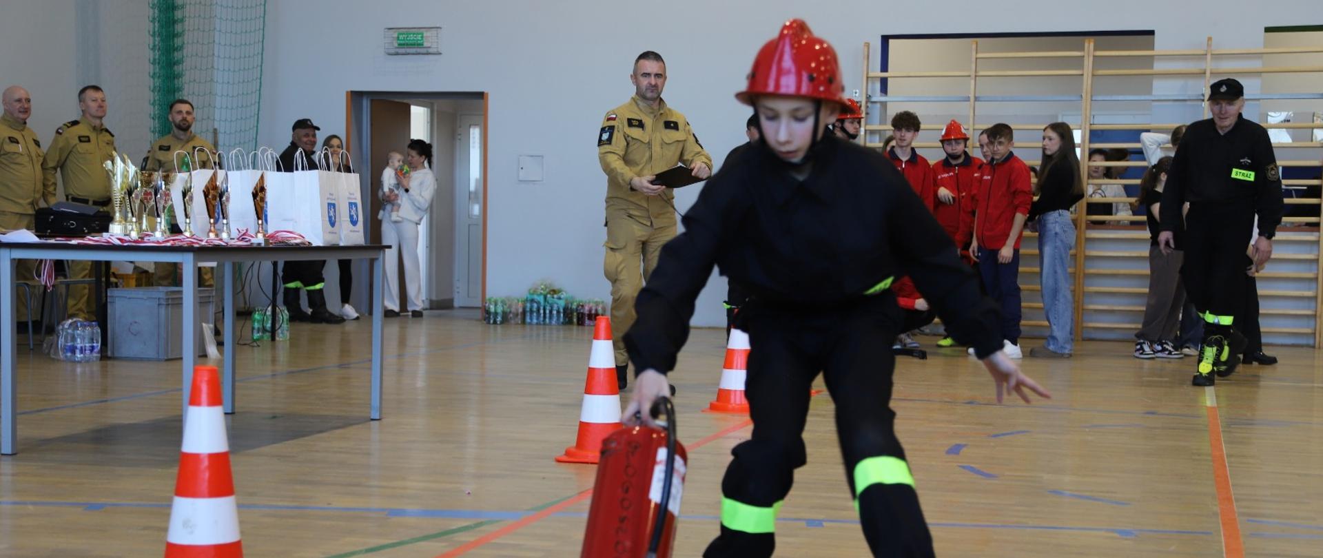 Zawodniczka podczas konkurencji ustawiania gaśnicy w odpowiednim miejscu. W tle sędzia zawodów oraz inni uczestnicy.