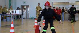 Zawodniczka podczas konkurencji ustawiania gaśnicy w odpowiednim miejscu. W tle sędzia zawodów oraz inni uczestnicy.