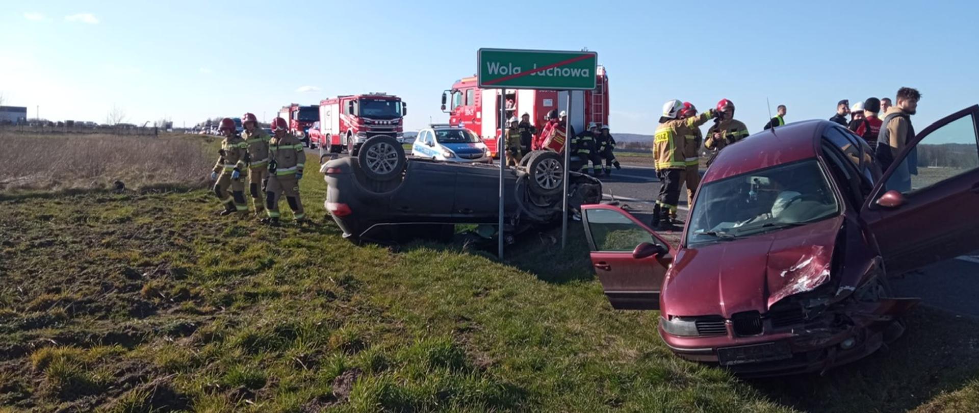 Zdjęcie przedstawia dwa z pośród czterech aut biorących udział kolizji drogowej. Jeden leży na dachu nad przydrożnym rowem, a drugi znajduje się w rowie. W głębi widać strażaków i trzy samochody pożarnicze. 