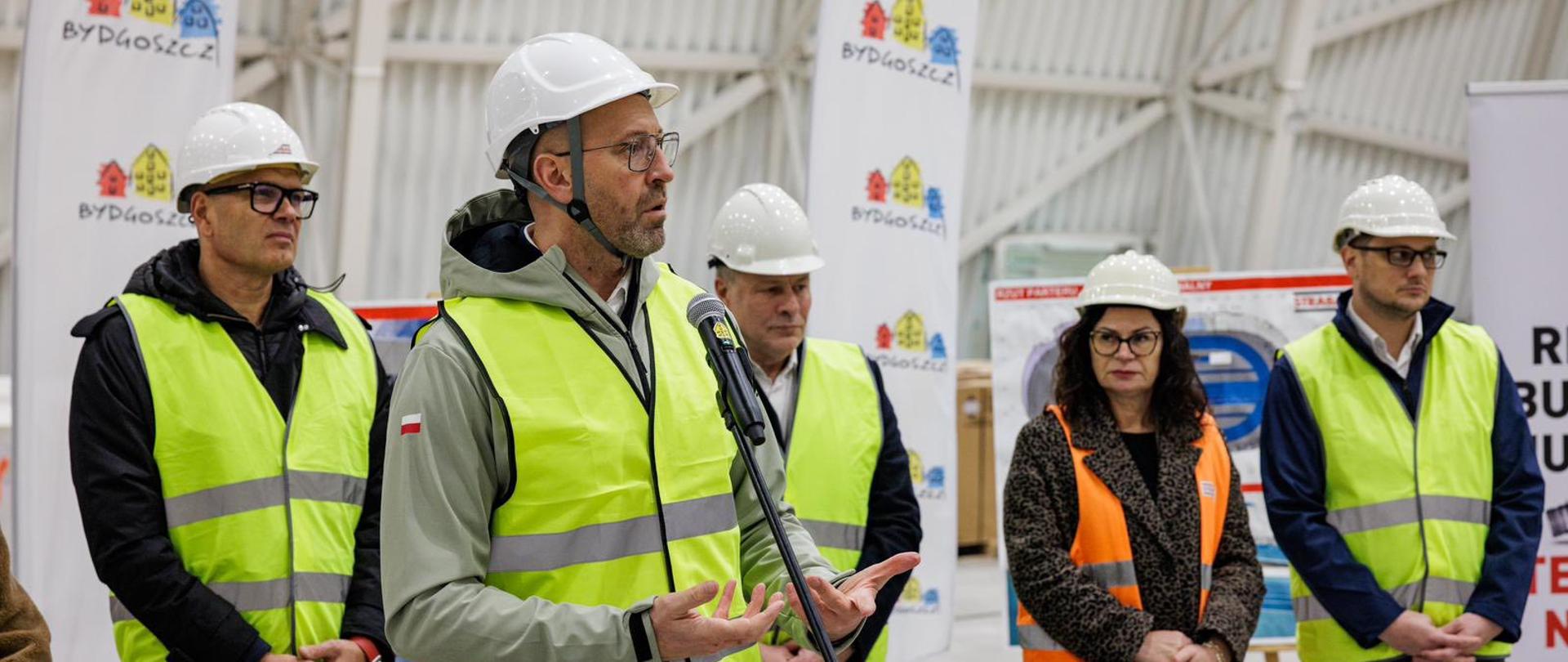 Minister sportu i turystyki Jakub Rutnicki w kasku i kamizelce odblaskowej mówi do mikrofonu podczas konferencji na budowanej hali sportowej. Za nim inni uczestnicy spotkania. Z tyłu windery z napisami "Bydgoszcz" i logiem miasta.