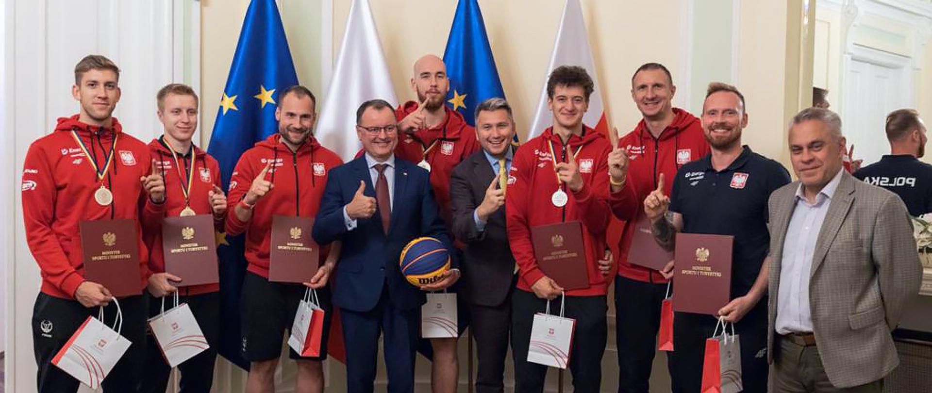 Spotkanie sekretarza stanu Arkadiusza Czartoryskiego z mistrzami świata - koszykarzami 3x3 U23 - zdjęcie zbiorowe wiceministra z koszykarzami na tle flag polskich i UE