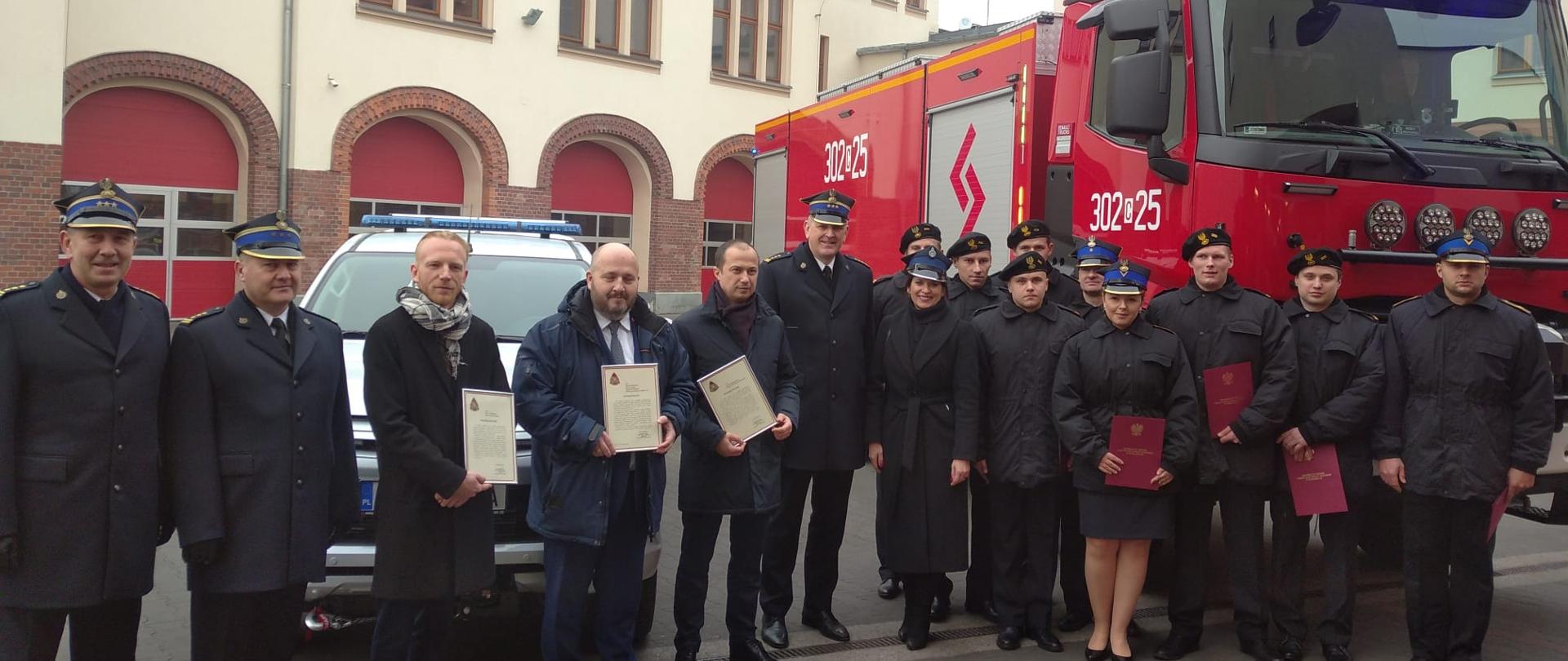 Stojący w rzędzie strażacy i oficjele w mundurach wyjściowych w kurtkach i płaszczach na placu z kostki JRG1 Bydgoszcz, za nimi dwa przekazywane pojazdy, z lewej biały terenowy Mitsubishi L200, z prawej Renault K440 ciężarowy koloru czerwonego