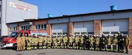 Strażacy stoją w rzędzie po egzaminie, ubrani w ubrania specjalne strażackie, w tle garaże JRG w Ostrołęce oraz samochód pożarniczy.