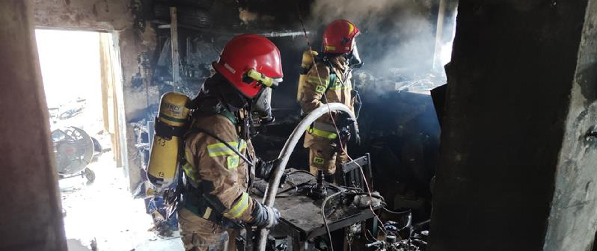 Dwóch strażaków ubranych w sprzęt ochrony osobistej strażaka trzyma linie gaśniczą polewając wodą wyposażenie spalonego garażu.