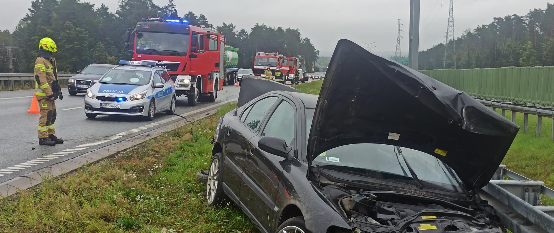Zdjęcie przedstawia uszkodzony samochód marki Volvo S60 z kompletnie rozbitym przodem. Samochód stoi na poboczu - po lewej widać strażaka oraz samochody policji i straży pożarnej. Na jezdni jest mokro.