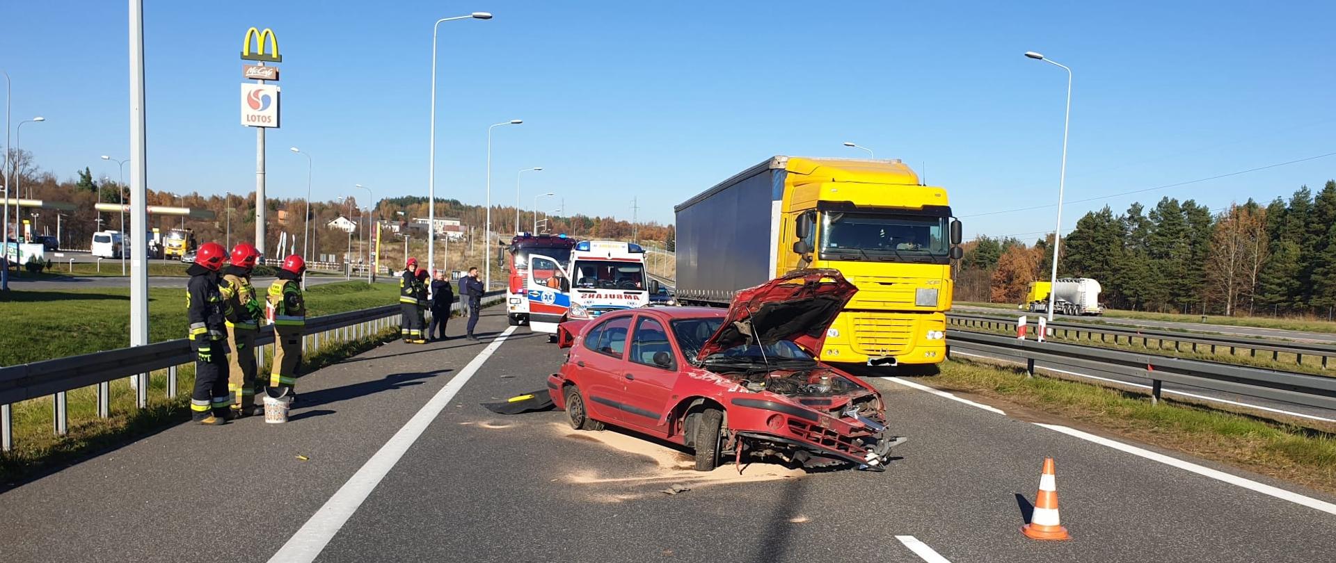 Na zdjęciu widać samochód osobowy, który uderzył w bariery energochłonne. Obok niego stoją strażacy ubrani w ubrania bojowe, pod samochodem widać rozsypany sorbent. Za samochodem jedzie ciężarówka i stoi ambulans wraz samochodem pożarniczym.