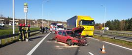 Na zdjęciu widać samochód osobowy, który uderzył w bariery energochłonne. Obok niego stoją strażacy ubrani w ubrania bojowe, pod samochodem widać rozsypany sorbent. Za samochodem jedzie ciężarówka i stoi ambulans wraz samochodem pożarniczym.