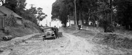Czarno-białe zdjęcie na którym znajduje się mężczyzna stojący tuż obok samochodu. Widoczna wyjeżdżona/ubita ziemia, która stanowi drogę. Po bokach drogi znajdują się domy.