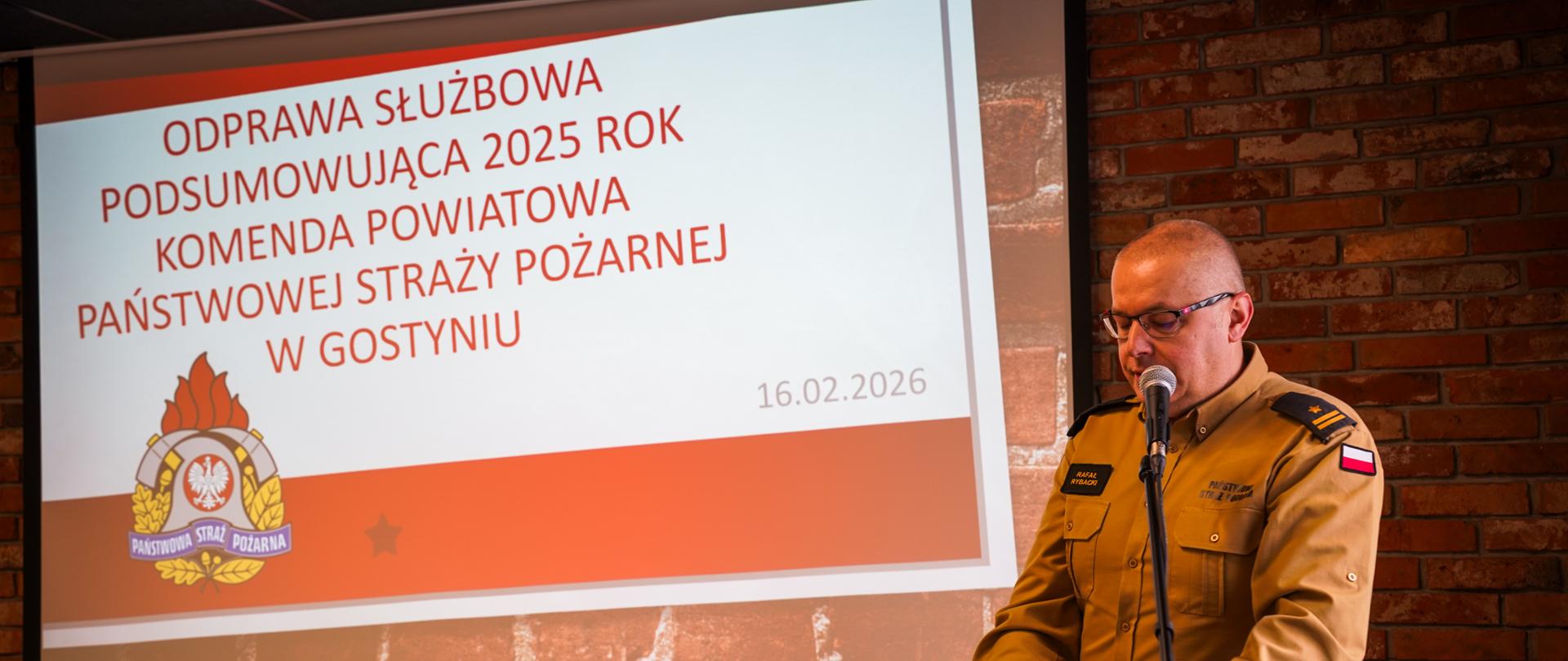 Komendanta Powiatowego, który stoi przy mównicy. Przedstawia on przybyłych gości na odprawę. W tle wyświetlana jest prezentacja.