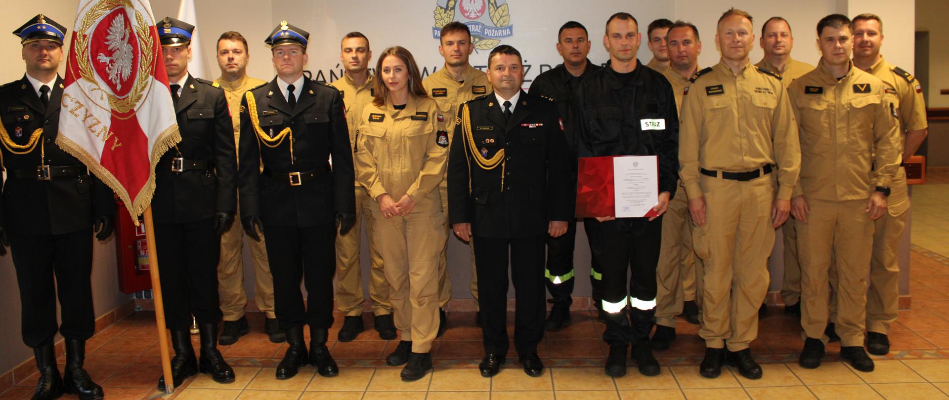 Uczestnicy uroczystej zbiórki stoją ustawieni w dwóch szeregach, funkcjonariusz, który złożył ślubowanie i trzyma w dłoniach teczkę koloru czerwonego.