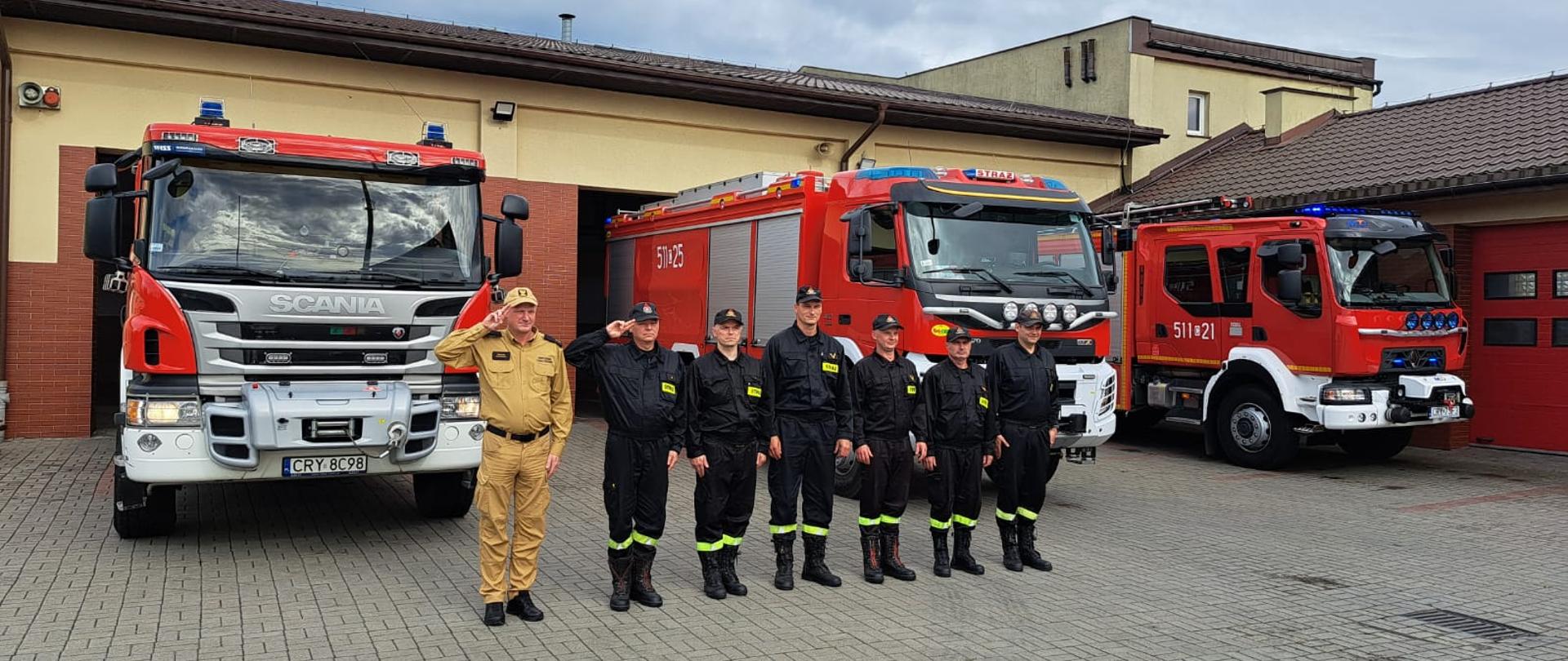 Siedmiu funkcjonariuszy Państwowej Straży Pożarnej w Rypinie wystąpiło na placu jednostki ratowniczo-gaśniczej w celu upamiętnienia siedemdziesiątej dziewiątej rocznicy Powstania Warszawskiego. Strażacy stoją w szeregu. Pierwszy od lewej strony jest Zastępca Komendanta Powiatowego Państwowej Straży Pożarnej w Rypinie - młodszy kapitan Marcin Czarnecki. Oddaje On honor, a wszyscy funkcjonariusze stoją na baczność. Za nimi znajdują się trzy samochody gaśnicze, a w tle garaże jednostki ratowniczo-gaśniczej.