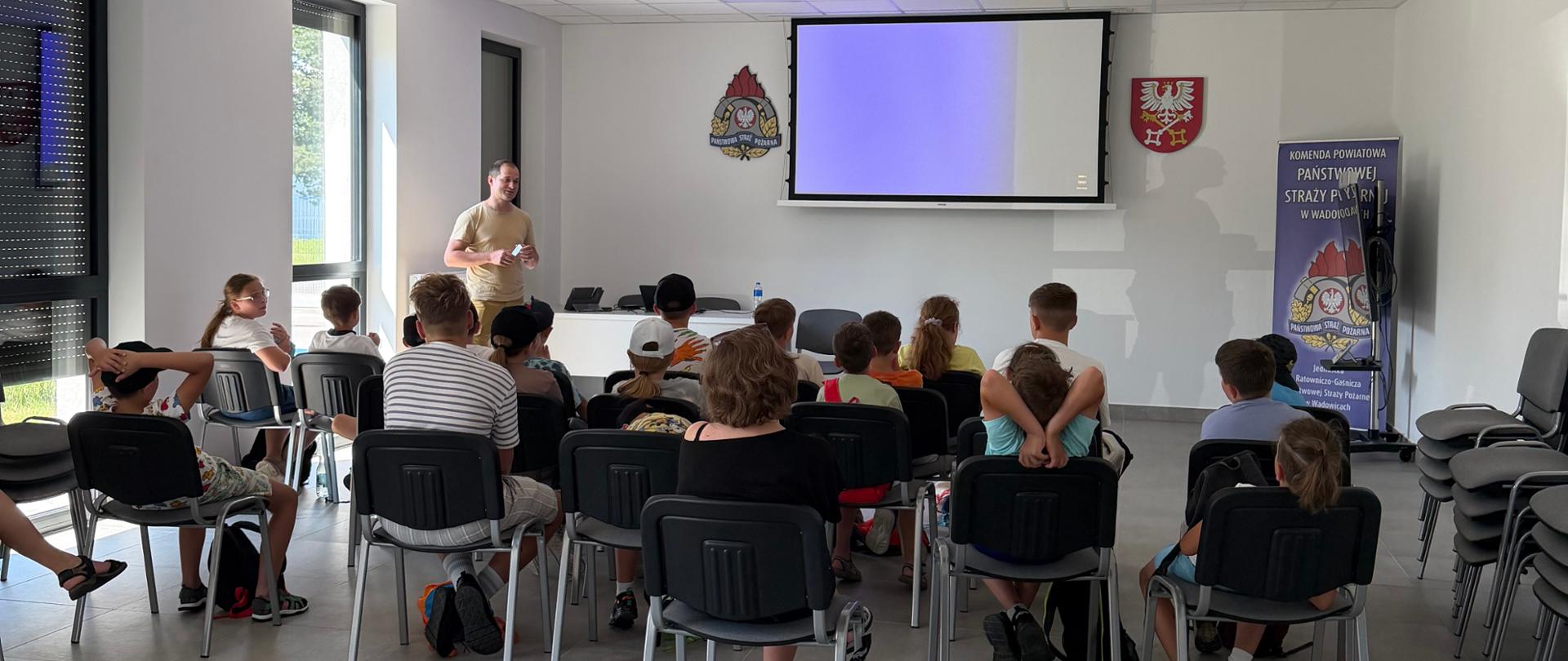 Na zdjęciu widać grupę dzieci przyglądającą się strażakowi który prowadzi zajęcia z CO i OL. Zajęcia są prowadzone w formie prezentacji multimedialnej. 