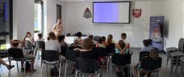 Na zdjęciu widać grupę dzieci przyglądającą się strażakowi który prowadzi zajęcia z CO i OL. Zajęcia są prowadzone w formie prezentacji multimedialnej. 