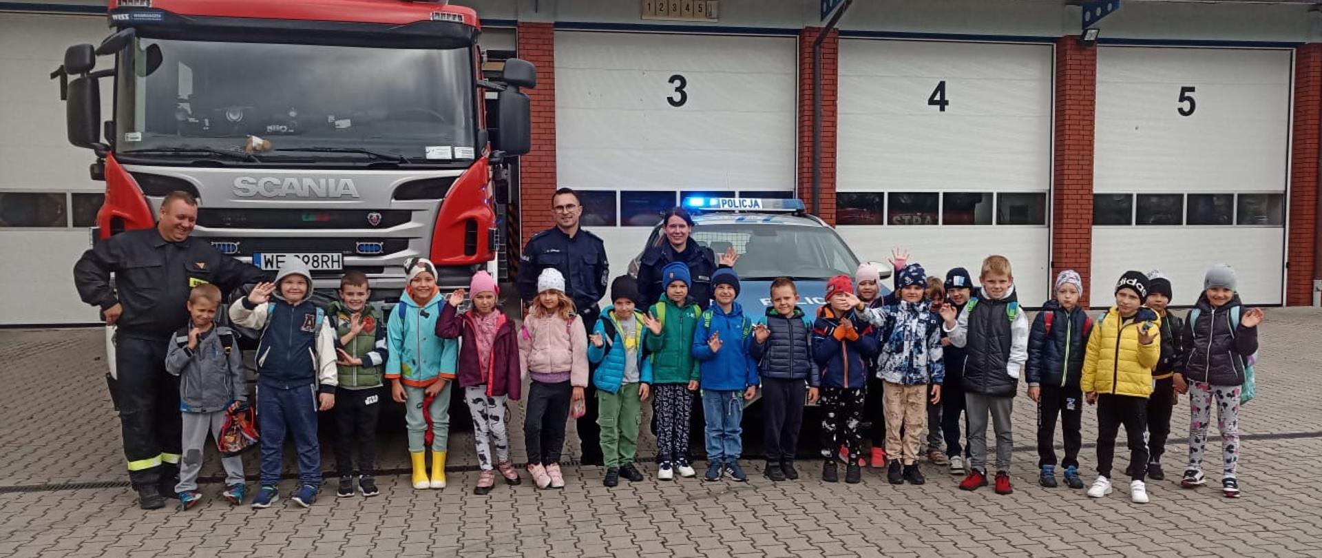 Dzieci stoją na tle garażu straży pożarnej pozując do wspólnego zdjęcia