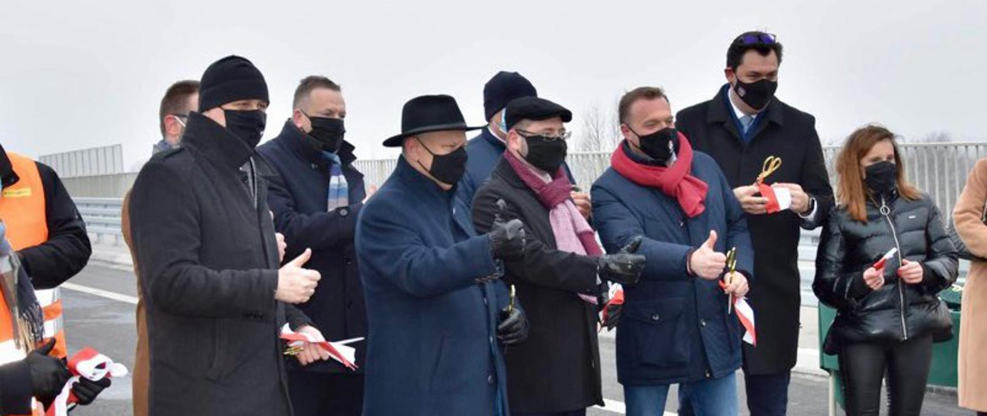 Elegancko ubrani ludzie biorą udział w uroczystości otwarcia wybudowanej drogi. Wszyscy pozują w kciukami uniesionymi do góry