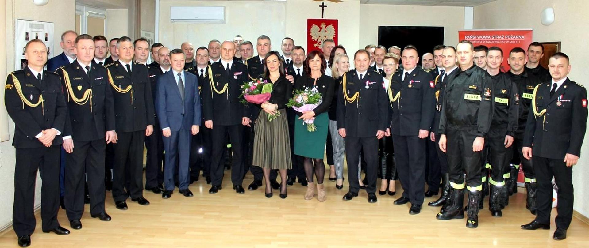 Fotografia przedstawia uroczysty apel na świetlicy komendy powiatowej Państwowej Straży Pożarnej w Mielcu. Fotografia przedstawia zdjęcie zbiorowe uczestników apel zorganizowanego na świetlicy komendy powiatowej Państwowej Straży Pożarnej w Mielcu. Na pamiątkowej fotografii uwiecznieni goście, obecni i byli funkcjonariusze, pracownicy cywilni z komendy powiatowej Państwowej Straży Pożarnej w Mielcu oraz najbliższa rodzina. Strażacy ubrani w mundury galowe koloru granatowego i mundury koszarowe.