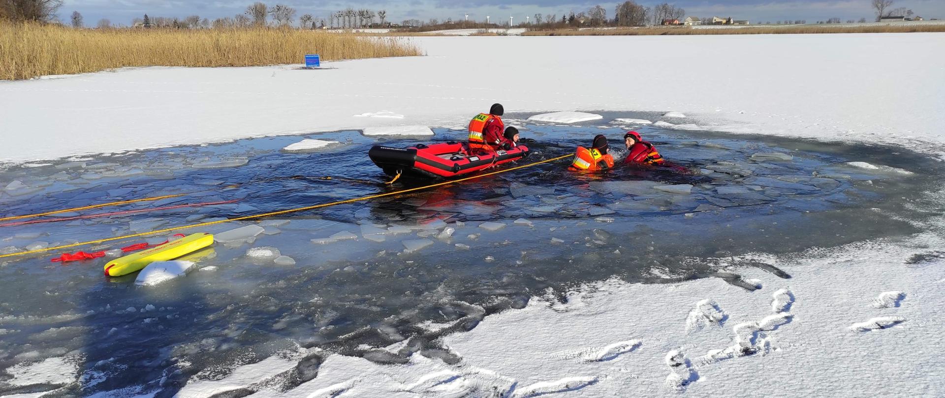 Zdjęcie przedstawia zamarznięte jezioro oraz dwóch strażaków będących w pontonie oraz 2 strażaków będących w wodzie. Sytuacja na zdjęciu przedstawia ćwiczenia strażaków, którzy pomagają osobie pod którą załamał się lód.
