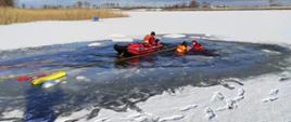 Zdjęcie przedstawia zamarznięte jezioro oraz dwóch strażaków będących w pontonie oraz 2 strażaków będących w wodzie. Sytuacja na zdjęciu przedstawia ćwiczenia strażaków, którzy pomagają osobie pod którą załamał się lód.