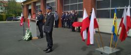 Na zdjęciu dwóch strażaków, jeden stoi na baczność, drugi przemawia za mównicą ubrani w mundurach wyjściowych, w tle bramy garażowe strażnicy, zaproszeni goście, flagi.