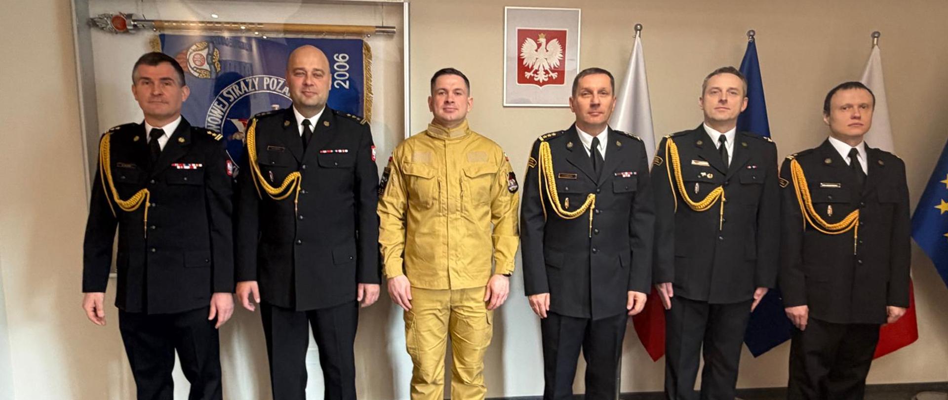 Grupowe zdjęcie sześciu mężczyzn w mundurach Państwowej Straży Pożarnej, stojących w rzędzie w oficjalnym pomieszczeniu. Pięciu mężczyzn ma na sobie czarne mundury wyjściowe ze złotymi sznurami galowymi na prawym ramieniu, baretkami odznaczeń na piersi oraz białymi koszulami i czarnymi krawatami. Trzeci mężczyzna od lewej wyróżnia się, mając na sobie piaskowy (beżowy) mundury służbowy.