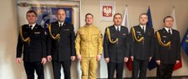Grupowe zdjęcie sześciu mężczyzn w mundurach Państwowej Straży Pożarnej, stojących w rzędzie w oficjalnym pomieszczeniu. Pięciu mężczyzn ma na sobie czarne mundury wyjściowe ze złotymi sznurami galowymi na prawym ramieniu, baretkami odznaczeń na piersi oraz białymi koszulami i czarnymi krawatami. Trzeci mężczyzna od lewej wyróżnia się, mając na sobie piaskowy (beżowy) mundury służbowy.