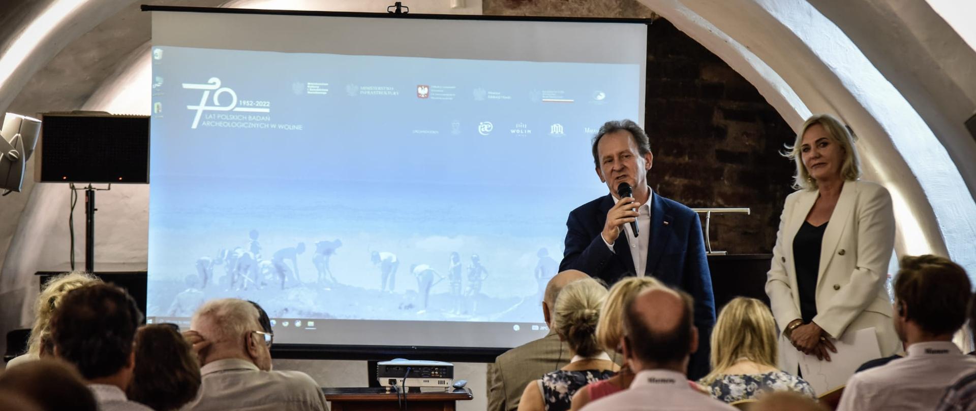 Mężczyzna stoi z mikrofonem i przemawia, obok stoi kobieta. Od tyłu widać ludzi uczestniczących w konferencji.