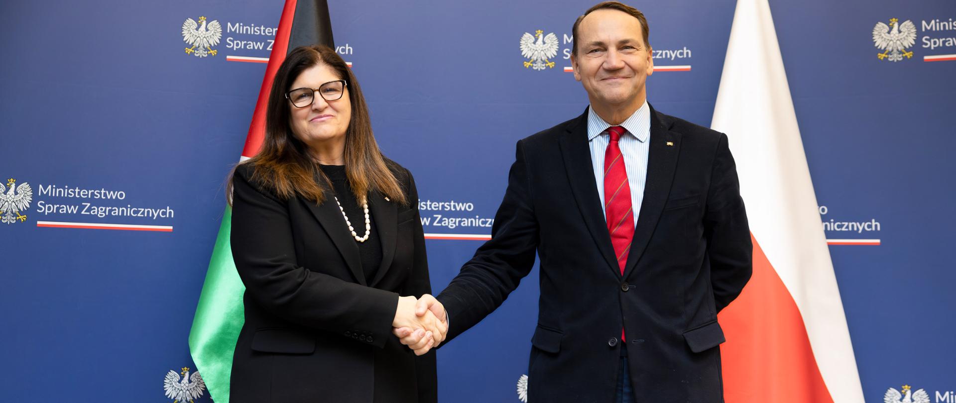 The Ministers of Foreign Affairs of Poland and Palestine stand in front of a backdrop with the Ministry of Foreign Affairs logo, with the flags of both countries visible behind them, and shake hands. Both are dressed in formal attire and face the camera while posing for an official photograph.