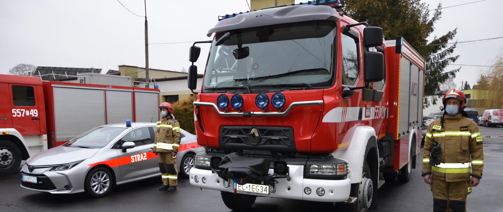Zdjęcie przedstawia dwa średnie samochody bojowe straży pożarnej i jeden samochód operacyjny stojący na placu przy Komendzie Powiatowej Państwowej Straży Pożarnej w Wieluniu, obok pojazdów stoją kierowcy w strojach bojowych