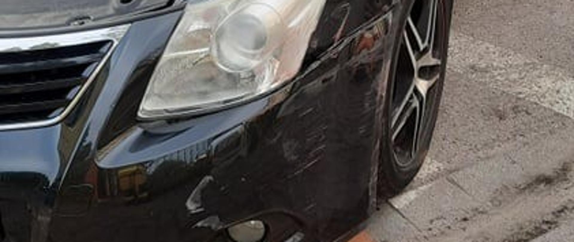 Zdjęcie przedstawia uszkodzony zderzak czarnego samochodu marki Toyota Avensis podczas kolizji na ul. Zamoyskiego w Białogardzie. Pojazd prezentowany od przodu z otwartą maskę silnika