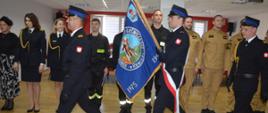 Świetlica komendy powiatowej. Pomieszczenie opuszcza poczet sztandarowy, który uczestniczył w ceremoni ślubowania. W tle, w szeregu stoją strażacy i pracownicy cywilni biorący udział w uroczystości.