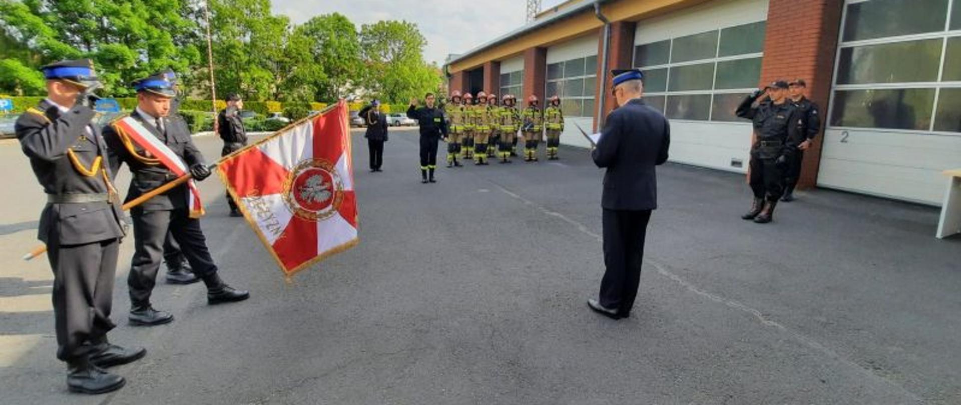 Na zdjęciu plac wewnętrzny strażnicy przed garażami. Po lewej stronie zdjęcia poczet sztandarowy, strażacy w ubraniach wyjściowych, w tyle kilkunastu strażaków ubranych w ubrania specjalne koloru beżowego i czerwone hełmy. Po środku zdjęcia Komendant w mundurze wyjściowym oraz strażak w ubraniu koszarowym składający ślubowanie.