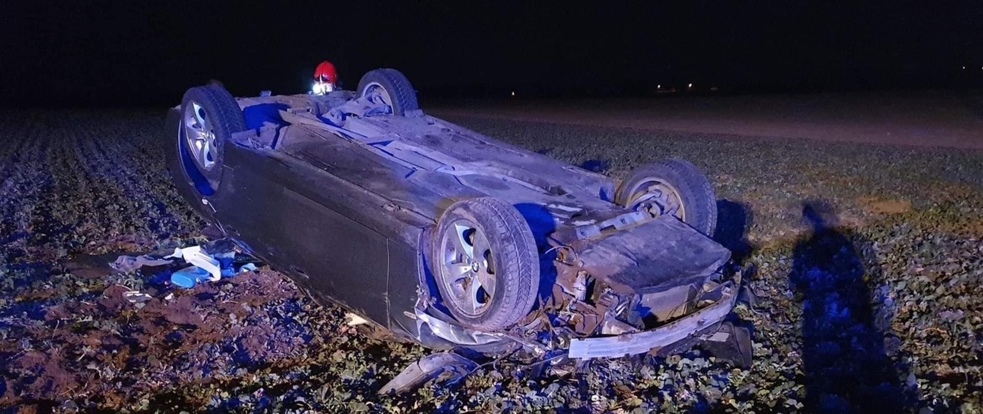 Samochód osobowy koloru ciemnego leży na dachu na polu. W prawym dolnym narożniku logo z napisem "KSRG Dąbrowa"