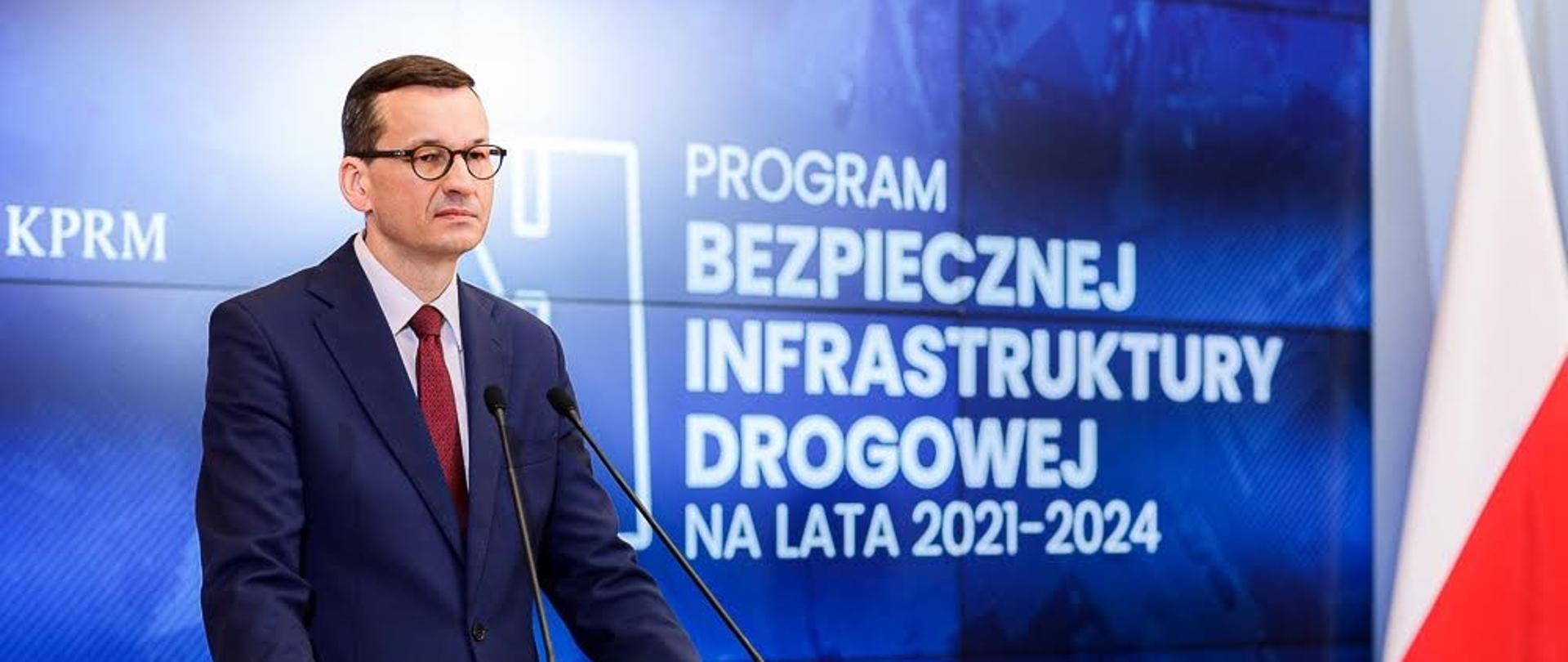 Premier Mateusz Morawiecki stoi przy mównicy. W tle flaga Polski oraz ekran z wyświetlaną prezentacją.