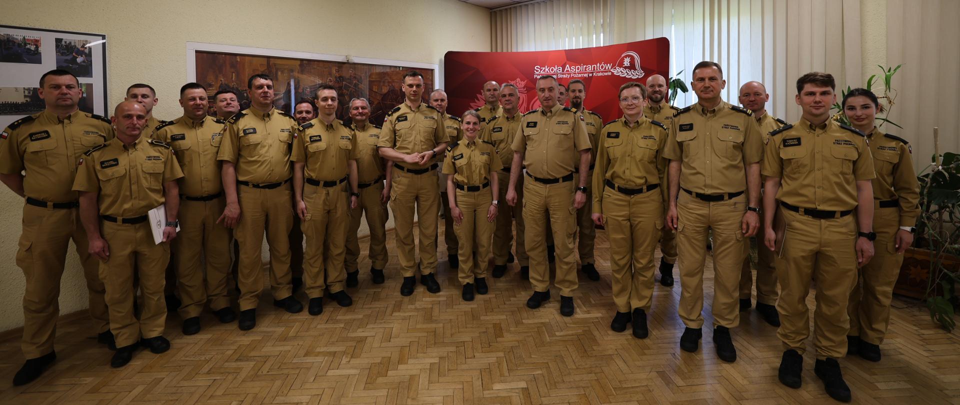 1. Na zdjęciu widoczna jest grupa około 25 funkcjonariuszy Państwowej Straży Pożarnej ubranych w jasnobrązowe (piaskowe) mundury służbowe. Osoby stoją w kilku rzędach w pomieszczeniu o jasnożółtych ścianach i jasnobrązowej podłodze z parkietu. W tle, po prawej stronie, znajduje się ścianka z logo Szkoły Aspirantów PSP w Krakowie, a za nią pionowe żaluzje na dużym oknie. Po lewej stronie na ścianie widoczne są zdjęcia i obrazy związane z tematyką strażacką. W centrum zdjęcia znajduje się Komendant Szkoły, nadbryg. Piotr Filipek, który wręcza dokument jednej z funkcjonariuszek. Obok niego stoi jego zastępca oraz pozostali funkcjonariusze – część z nich uśmiechnięta, wszyscy patrzą w stronę obiektywu. Zdjęcie ma charakter oficjalny, pamiątkowy.