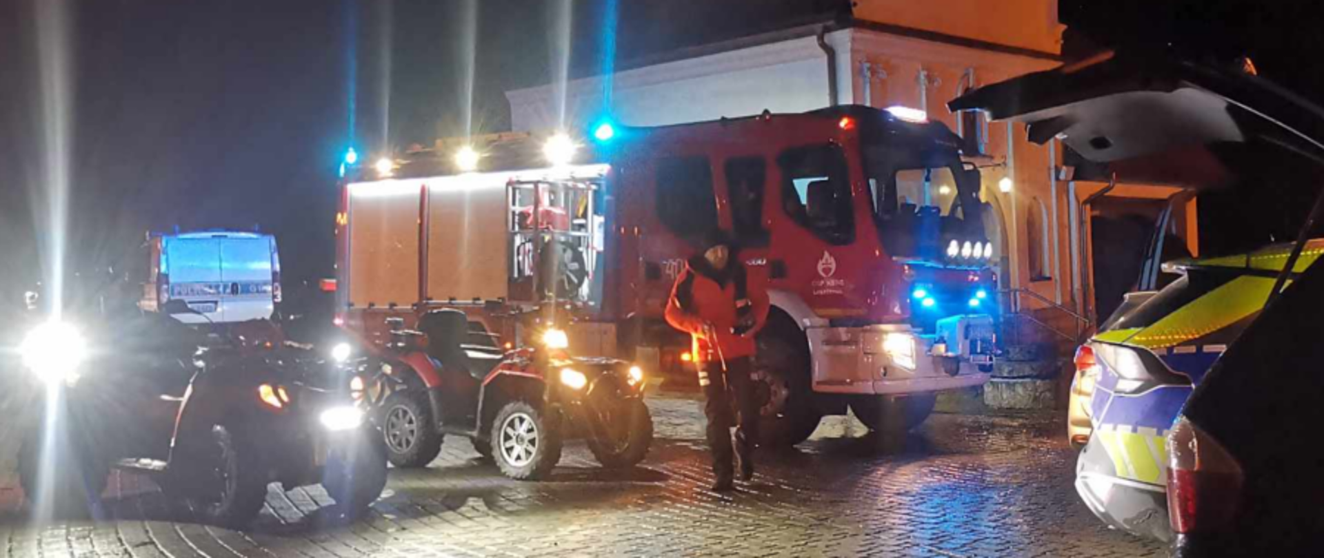 Zdjęcie zostało wykonane w porze nocnej na placu obok kaplicy, przedstawia wóz strażacki Volvo GBARt należący do OSP KSRG Leszczyna oraz dwa quady jeden również należący do tej jednostki zaś drugi do JRG Bochnia. Możemy również dostrzec mężczyznę obok tych pojazdów a także dwa radiowozy Policji, pierwszy znajduje się za pojazdami straży natomiast kolejny pomiędzy dwoma samochodami osobowymi w prawej części zdjęcia.