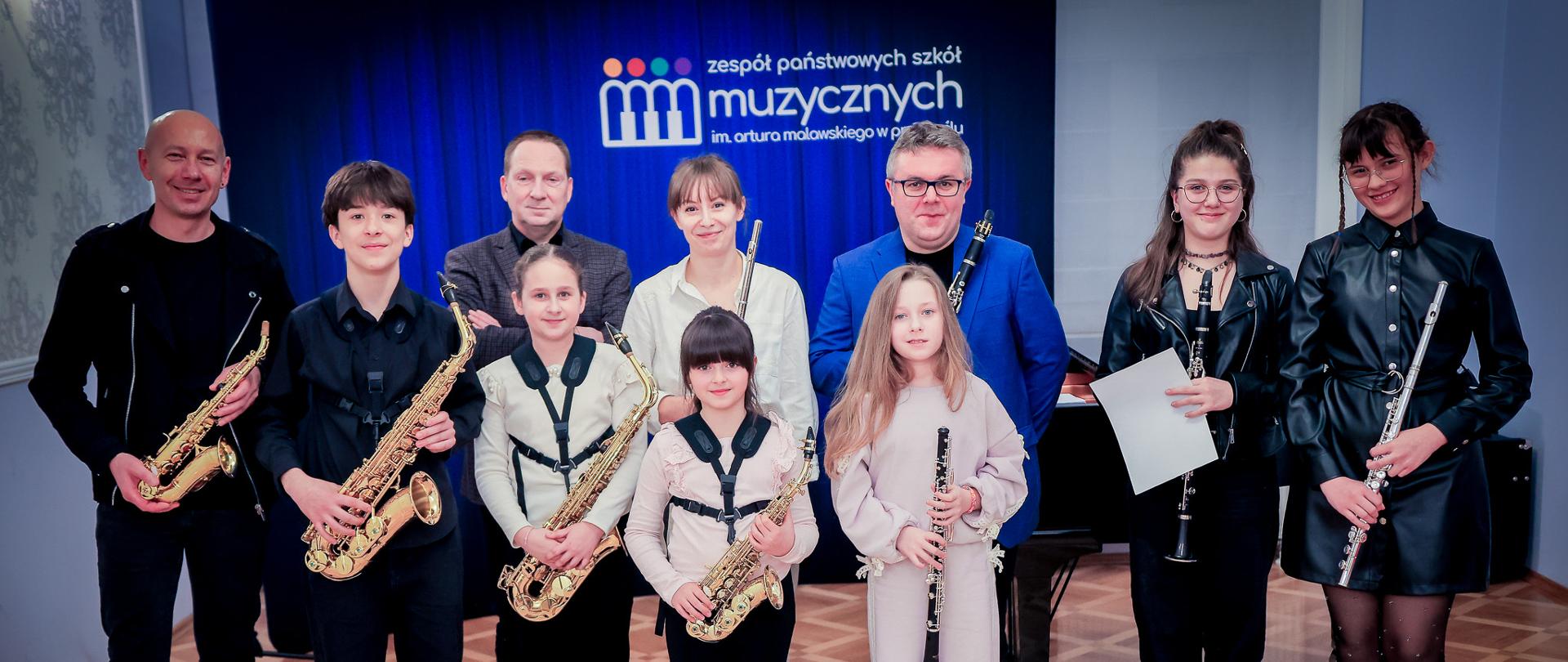 Grupa dzieci i dorosłych pozujących do zdjęcia w szkole muzycznej; uczestnicy trzymają instrumenty dęte, w tym saksofony, klarnety i flet, stojąc przed niebieskim tłem z logo szkoły, na drewnianej podłodze, obok fortepianu.